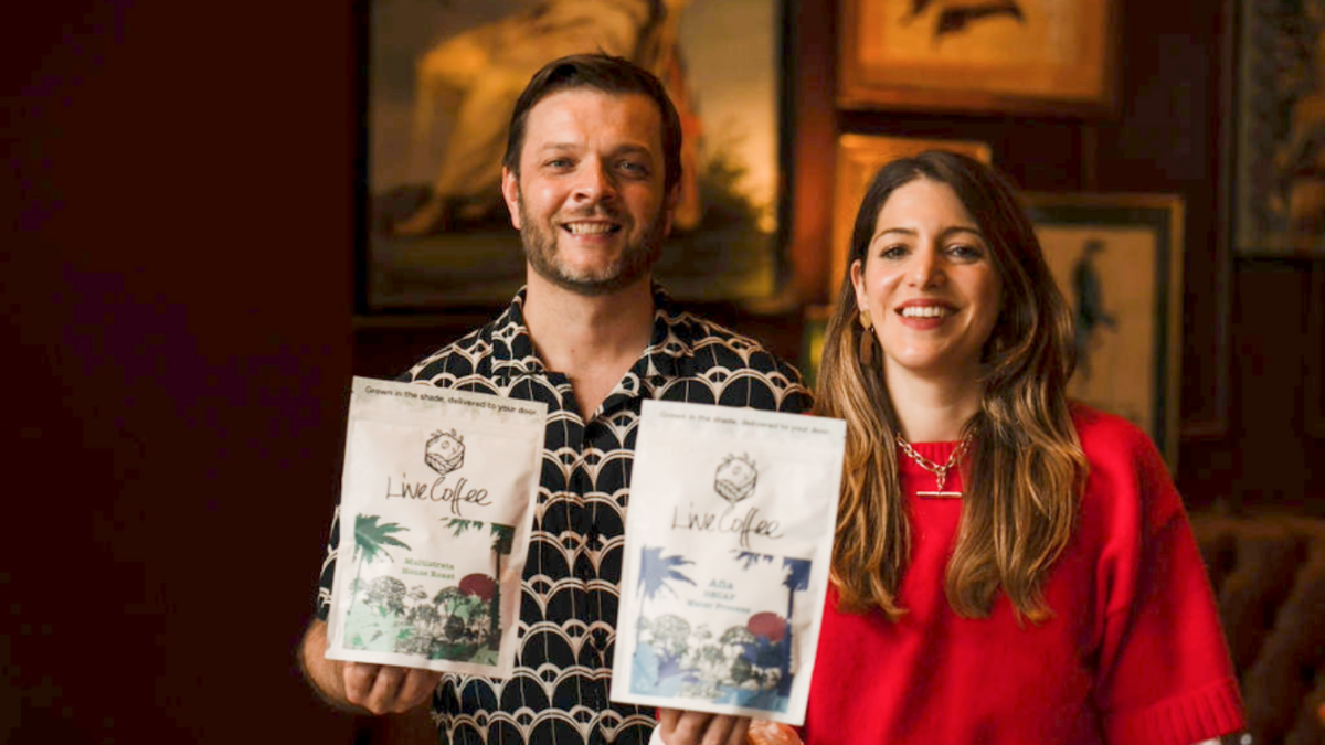 Founders Josh and Rosie standing indoors and holding two Live coffee bags. The man wears a black-and-white patterned shirt, and the woman wears a bright red top.